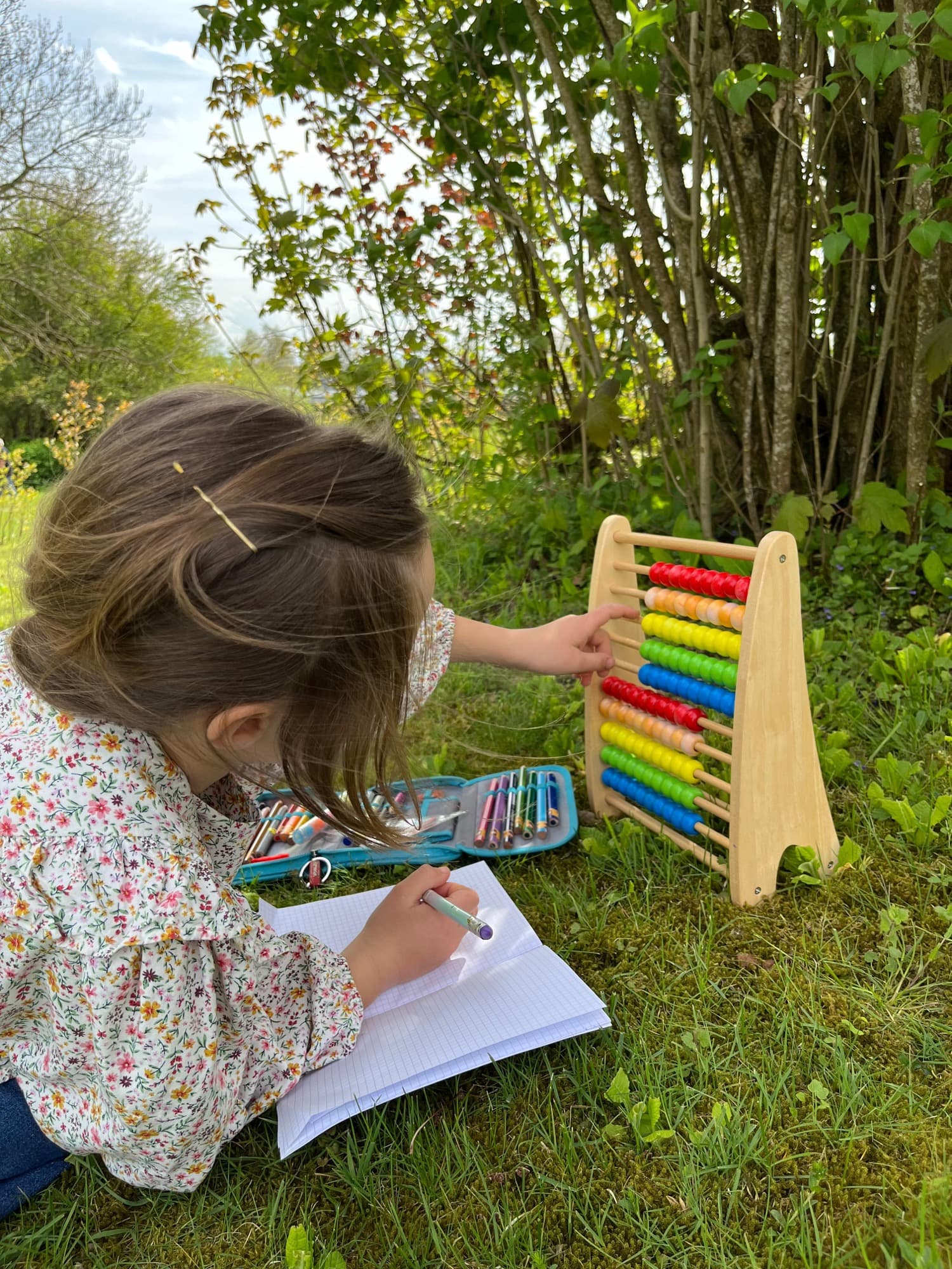 Mädchen sitzt auf einer Wiese, schreibt in ein Heft und rechnet an einem hölzernen Rechenschieber mit bunten Perlen.